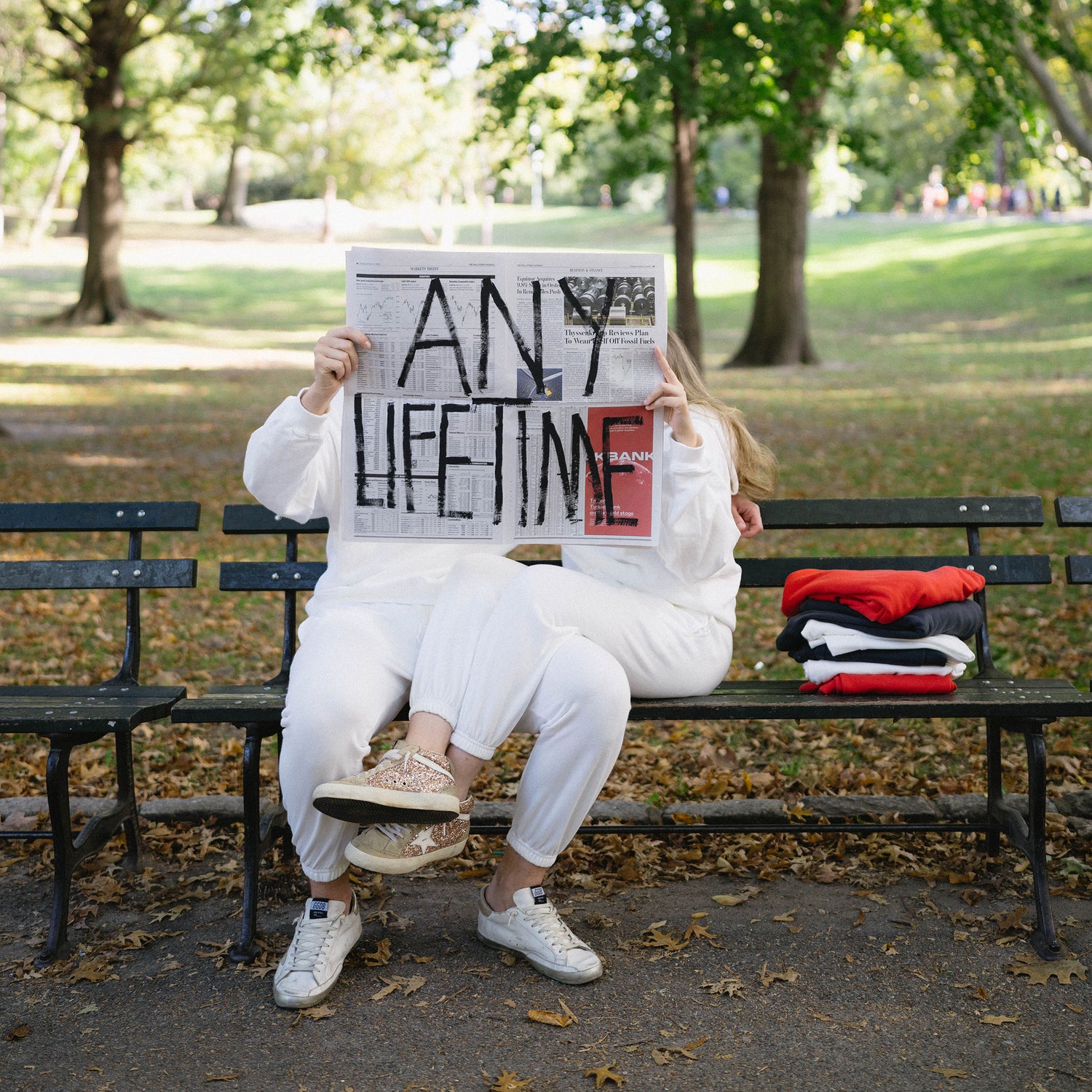 Miranda and Alex sitting on a bench reading a newspaper with "Any Lifetime" written on it in large letters.